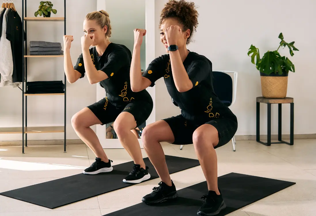 Snelle resultaten met EMS training in Mechelen Twee vrouwen in EMS-pakken doen squats en gebruiken EMS-training in Mechelen om in korte tijd snelle resultaten te boeken.