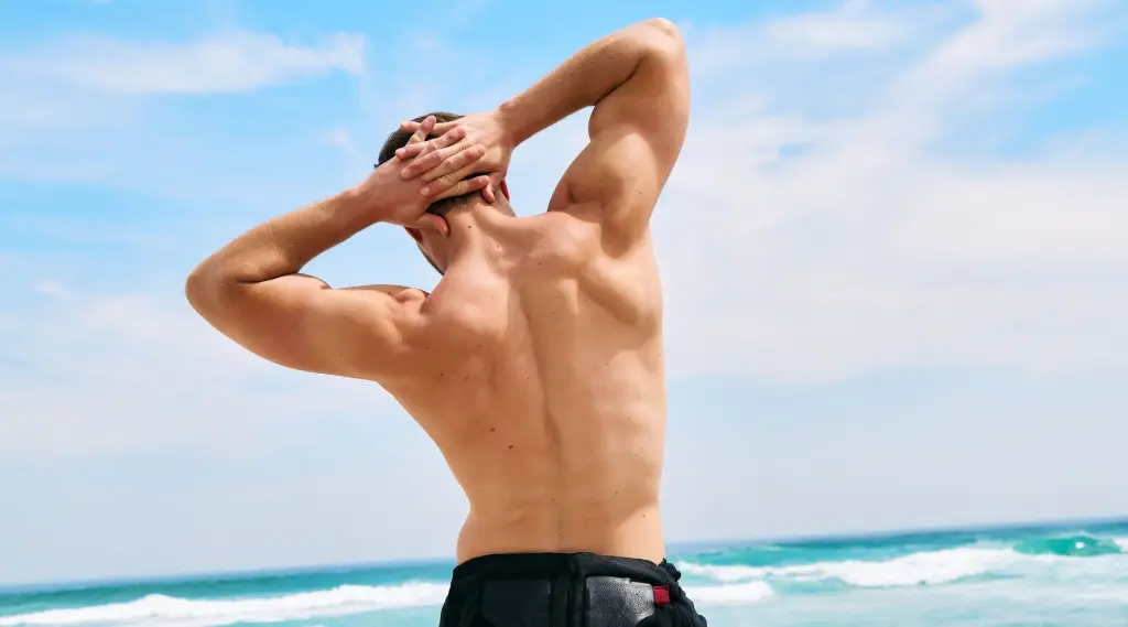 Un homme s'étire le dos sur la plage Un homme musclé vu de dos, plaçant ses mains derrière la tête tout en étirant son dos. Il se tient sur une plage, avec des vagues et un ciel bleu en arrière-plan. L’image met en valeur son dos fort et bien développé.
